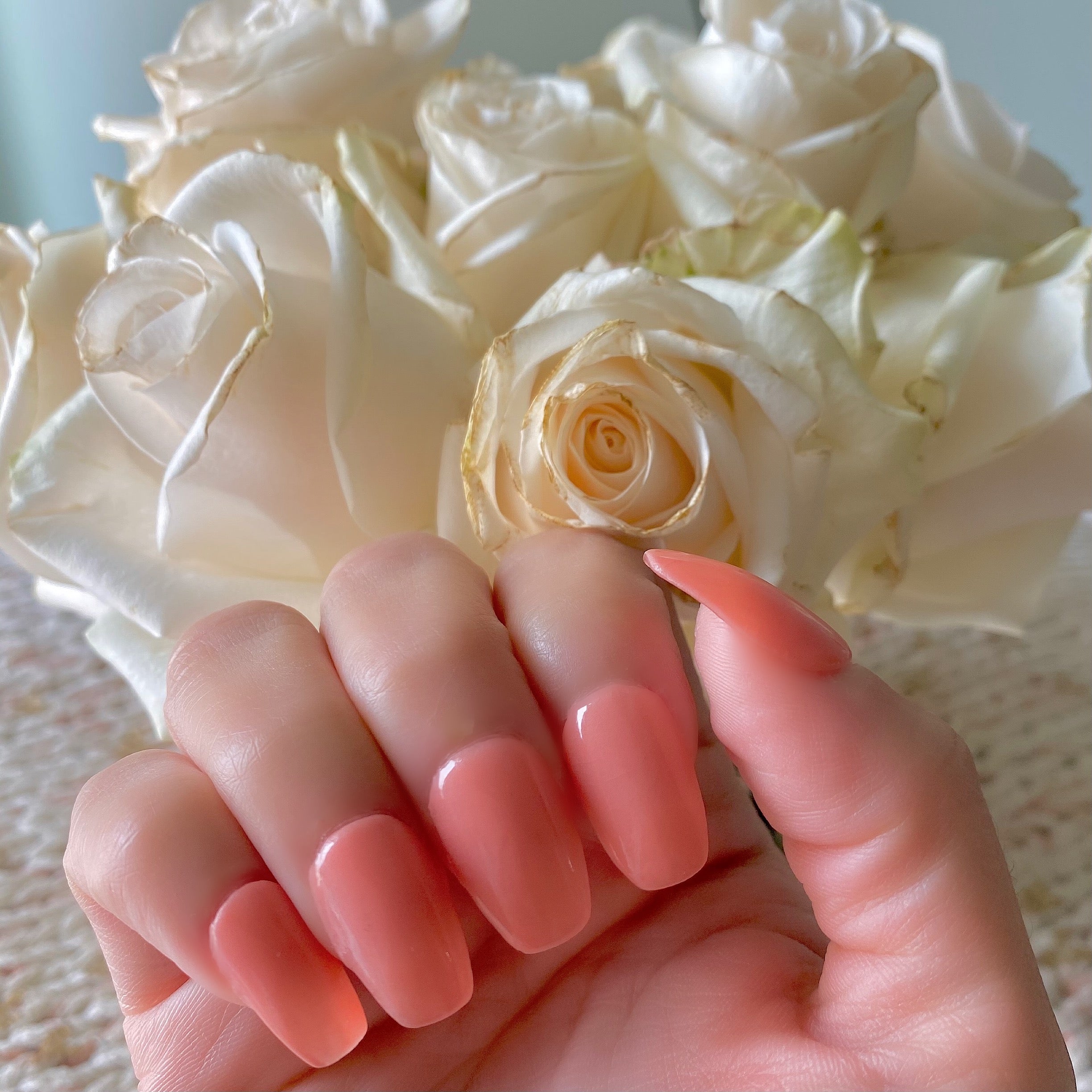 Long almond nails painted with solid peachy coral glow-in-the-dark dip powder over a backdrop of creamy white roses.