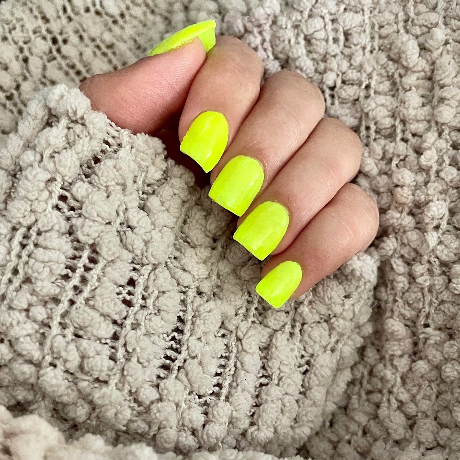Bright neon yellow-green square acrylic nails on hand against textured beige knit fabric.