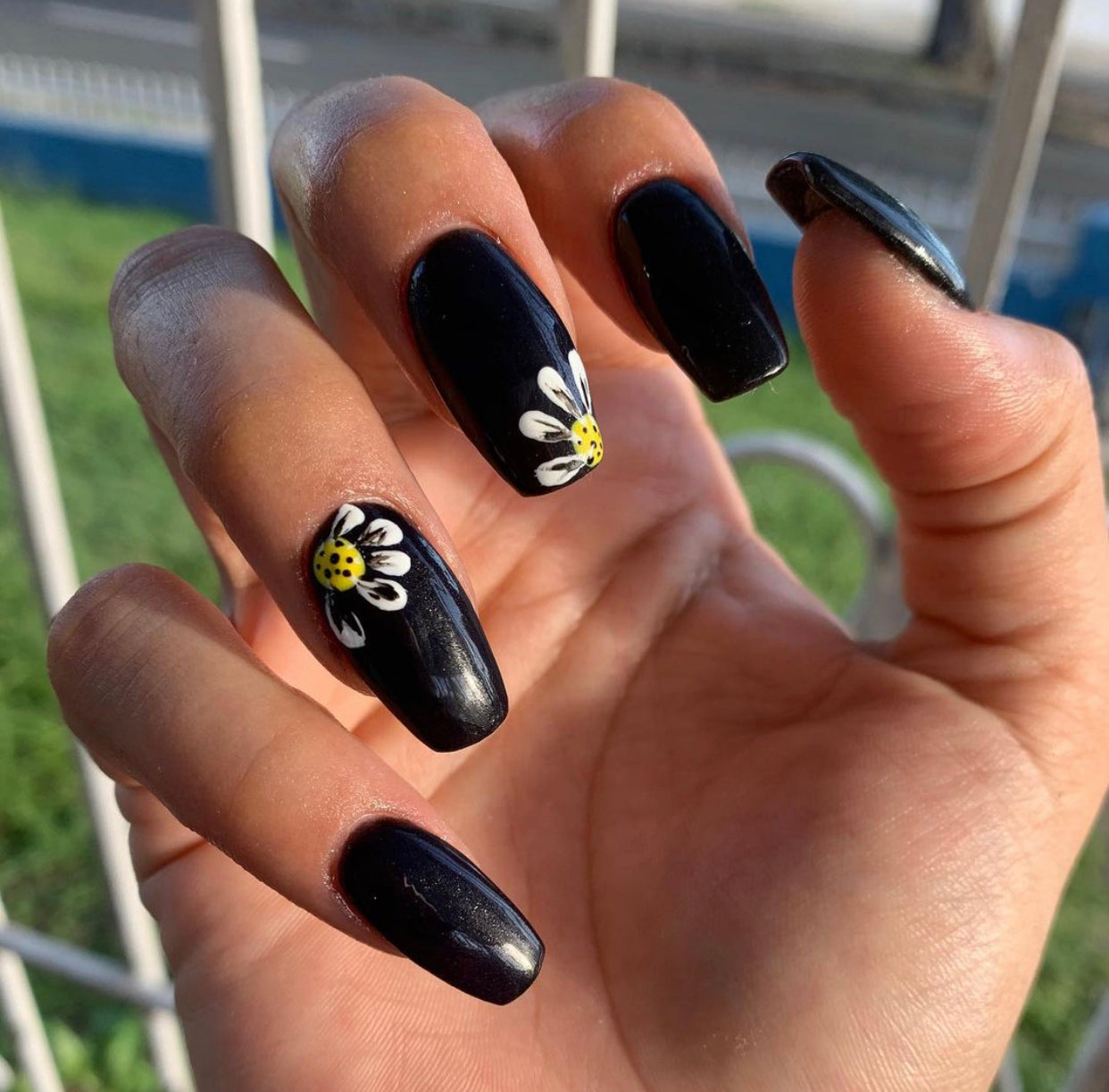 Glossy black square nails with hand-painted white and yellow daisy accents on ring and middle fingers.