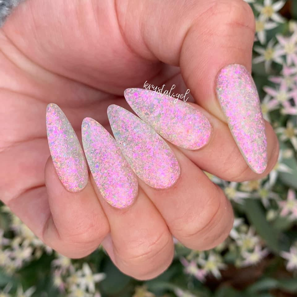 Long almond nails with pastel pink base and iridescent chameleon glitter flakes creating a shimmering fairy dust effect.
