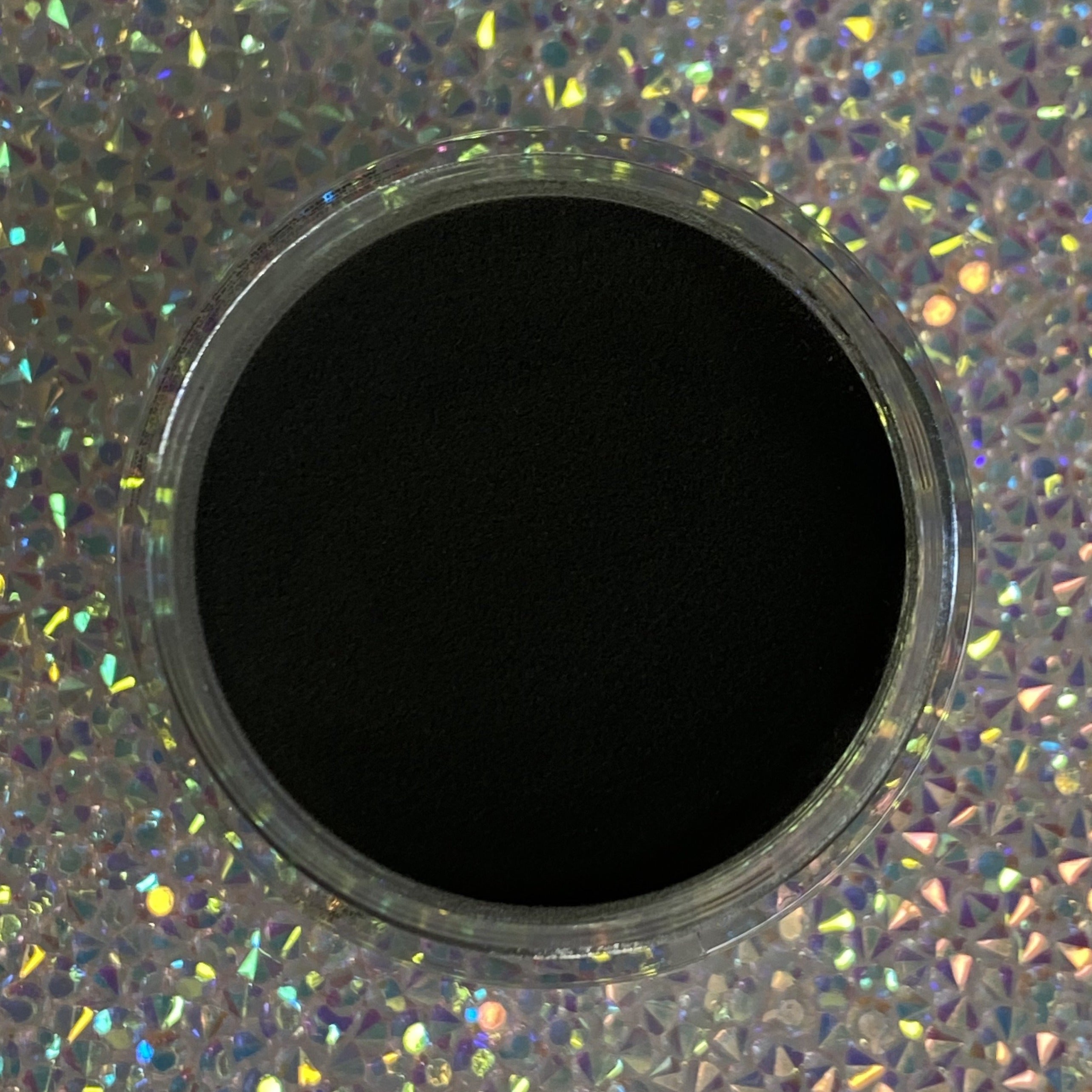 Open container of smooth, deep black dip powder on sparkly holographic surface.
