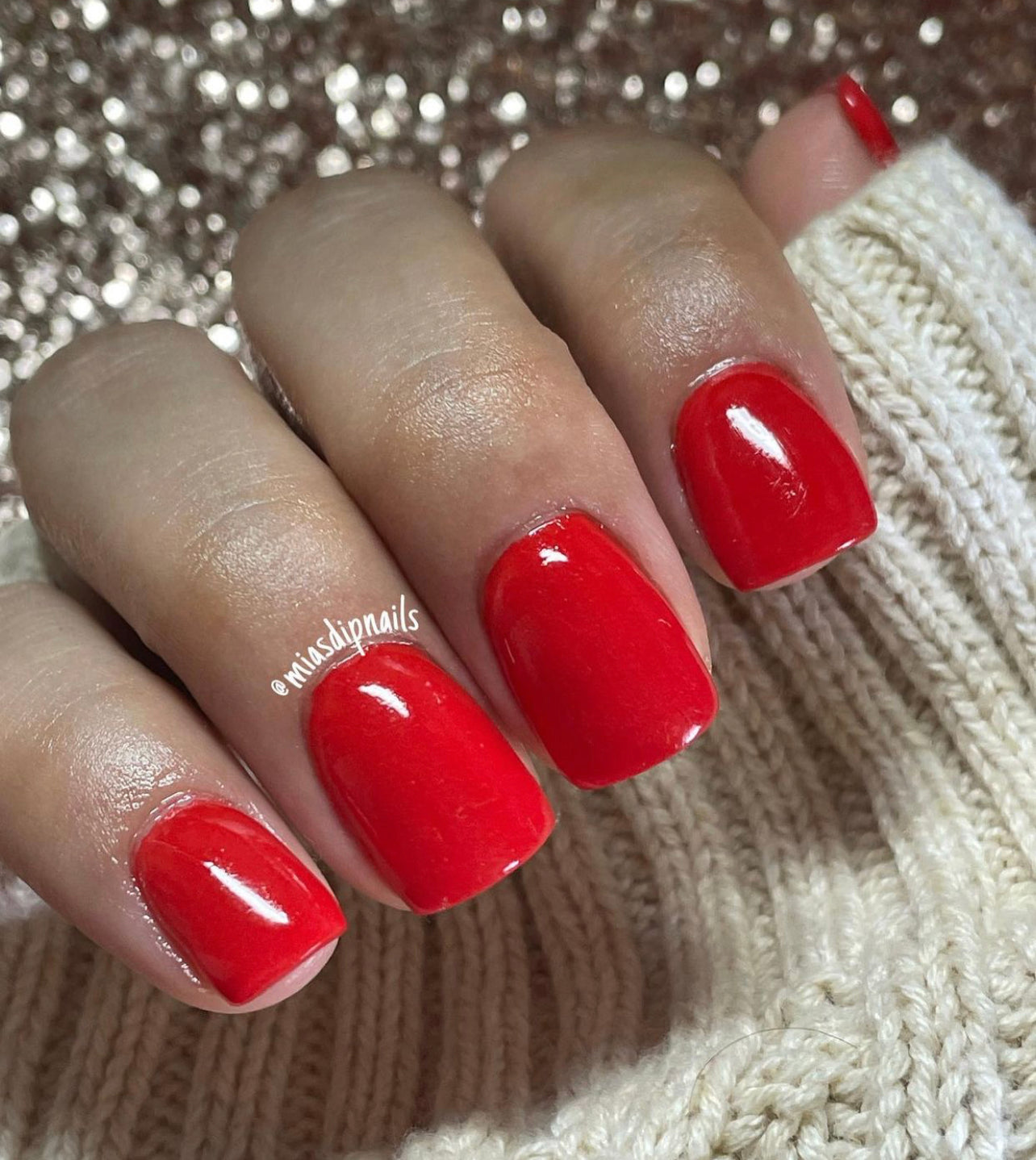 Bright glossy red dip powder nails on medium-length square tips with cream knit background.