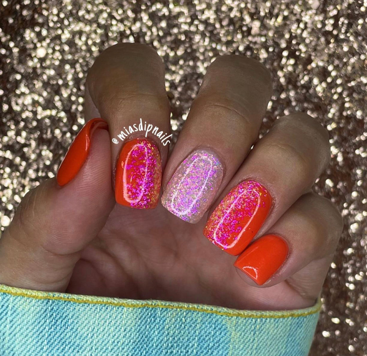 Bright orange dip powder nails with two accent nails featuring pink holographic glitter on a square shape.