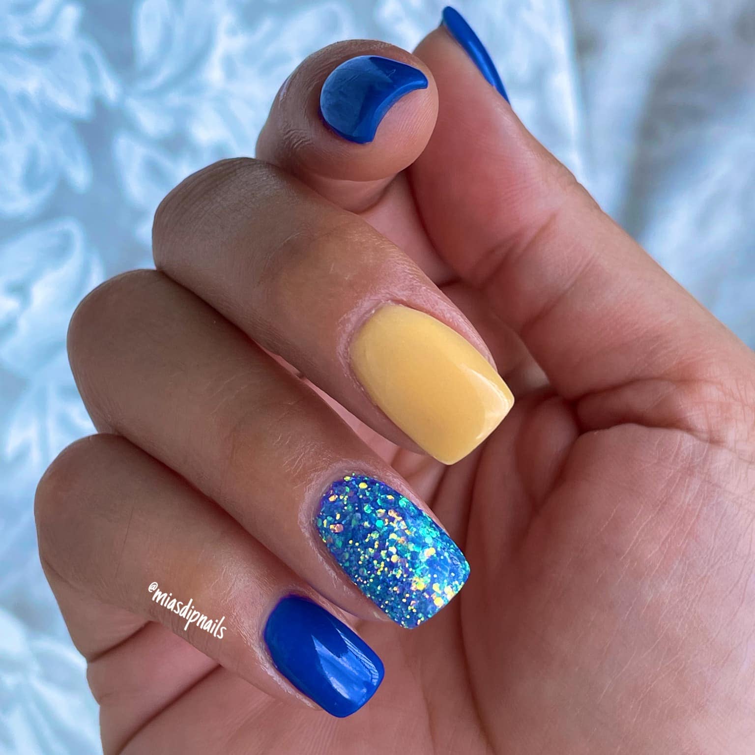 Hand with nails painted bright cobalt blue, soft pastel yellow, and glittery blue with iridescent flakes.