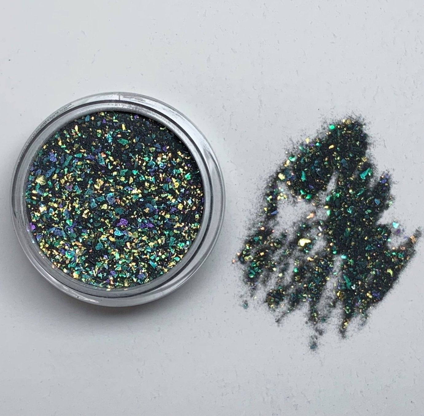 Small container of black dip powder with multicolor chameleon flakes scattered beside it on a white surface.