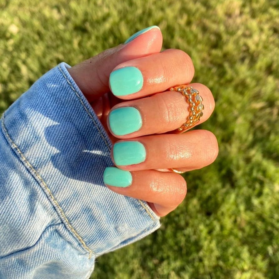 Teal solid dip powder nails on hand with gold chain rings and denim jacket sleeve outdoors.