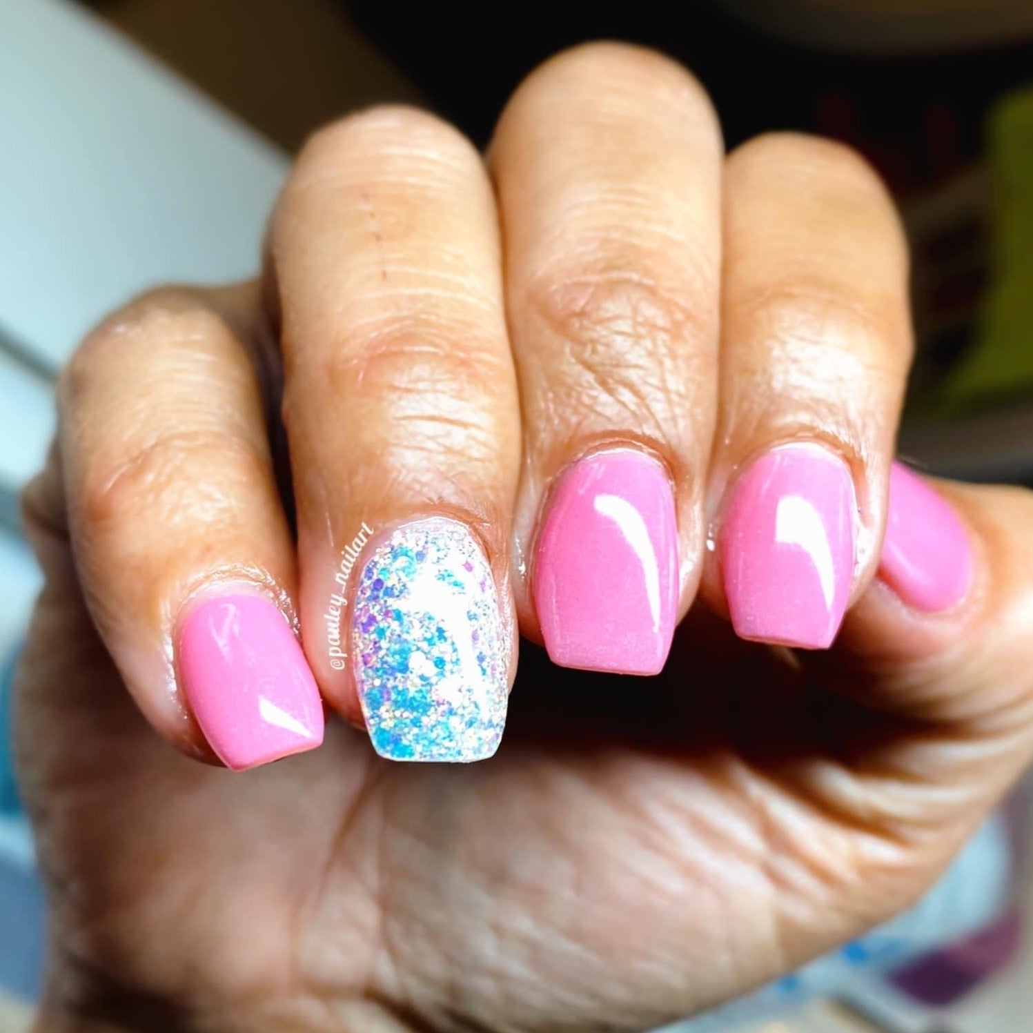 Hand with pink solid nails and white accent nail with blue glitter polish.