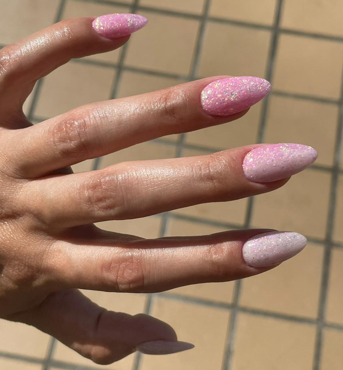 Long almond-shaped nails with pink and beige chameleon flake glitter dip powder design on hand against tiled background.