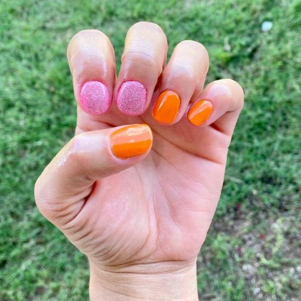 Hand with short nails coated in bright orange and textured pink dip powder polish against a blurred green grass background.