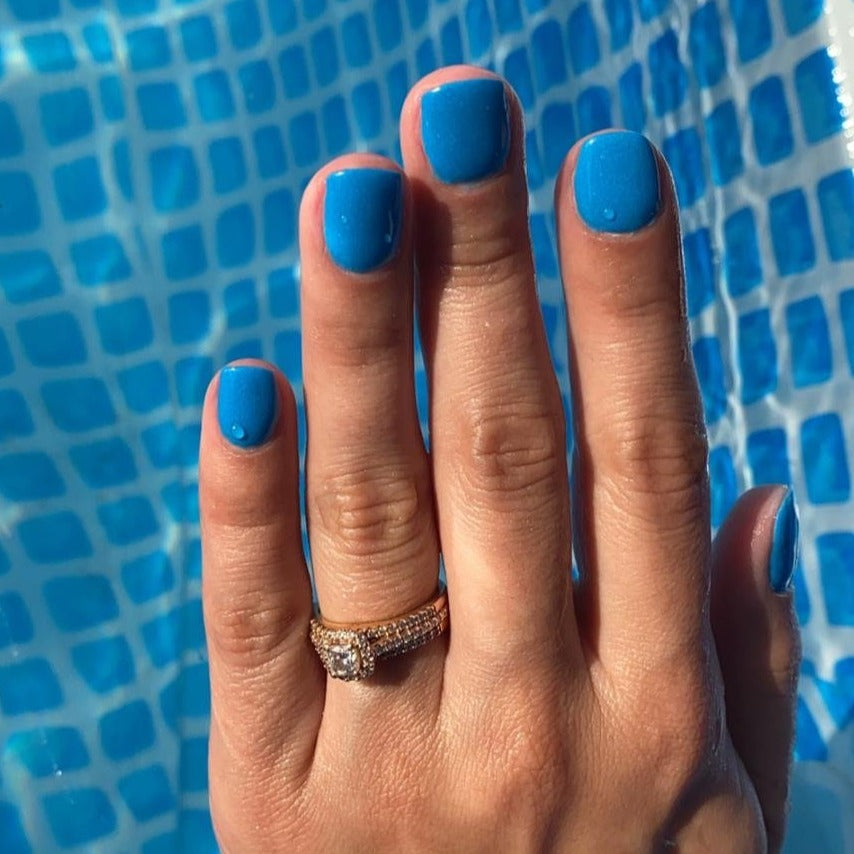 Bright neon blue dip powder nails on hand with gold and diamond ring over blue tiled pool background.