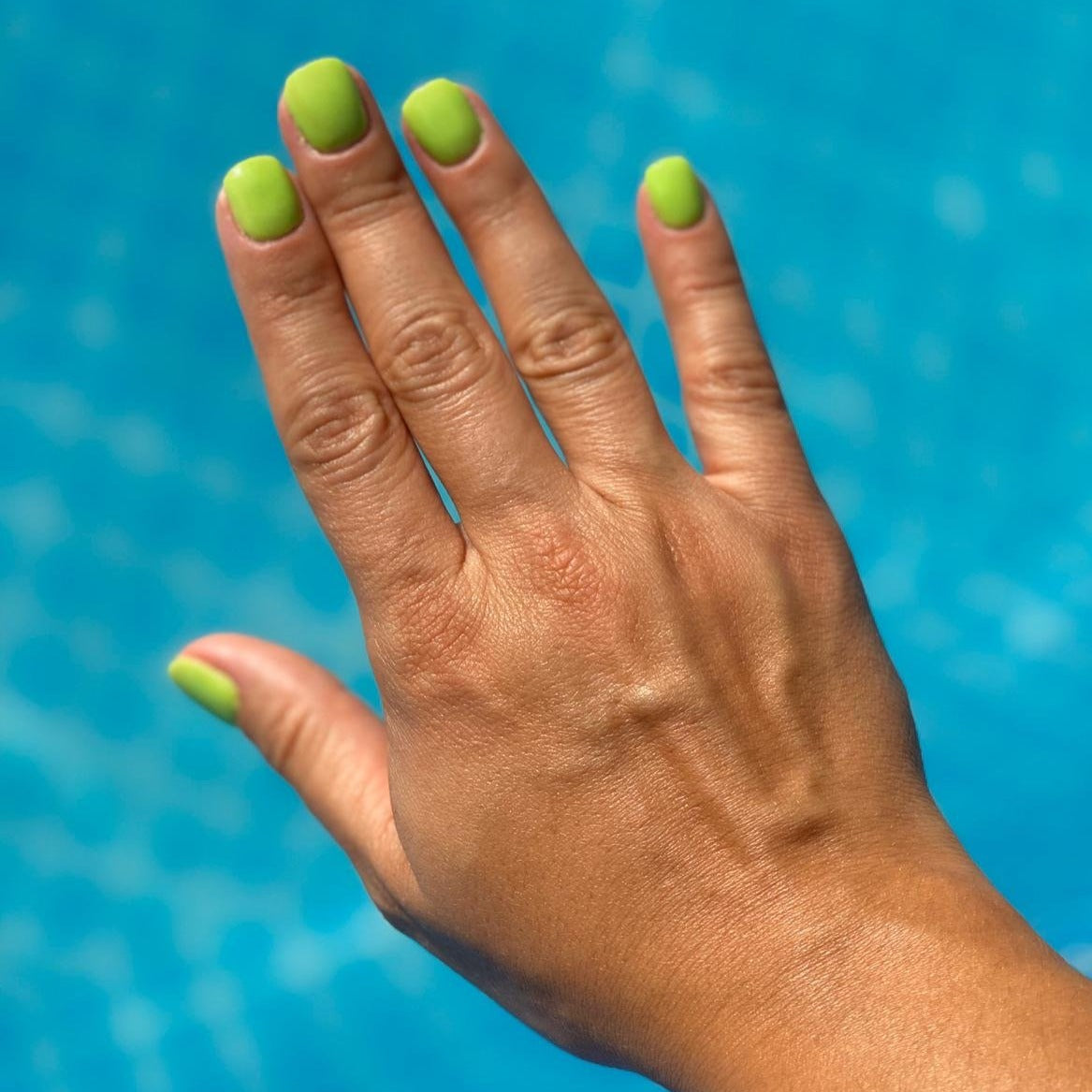Hand with matte neon green dip powder nails against a bright blue textured background.