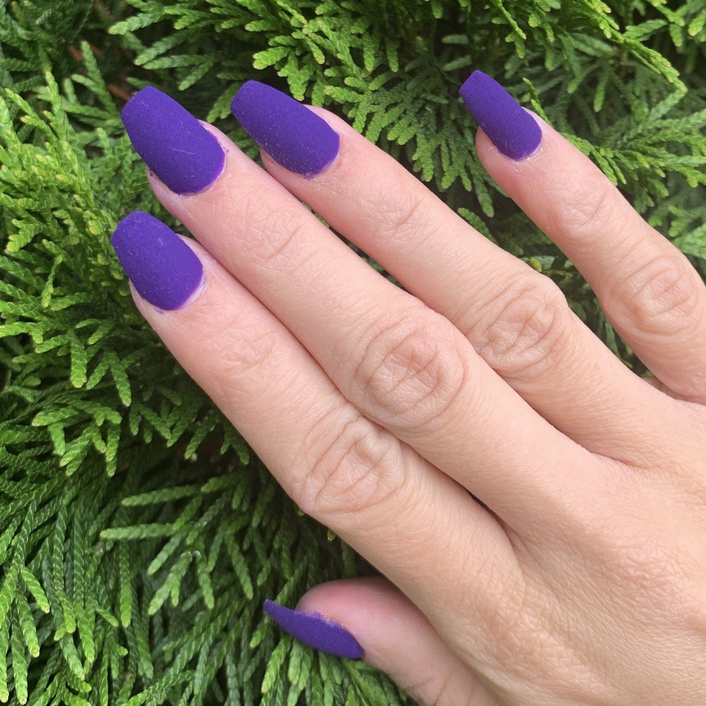 Matte purple coffin-shaped nails on hand against green foliage background.