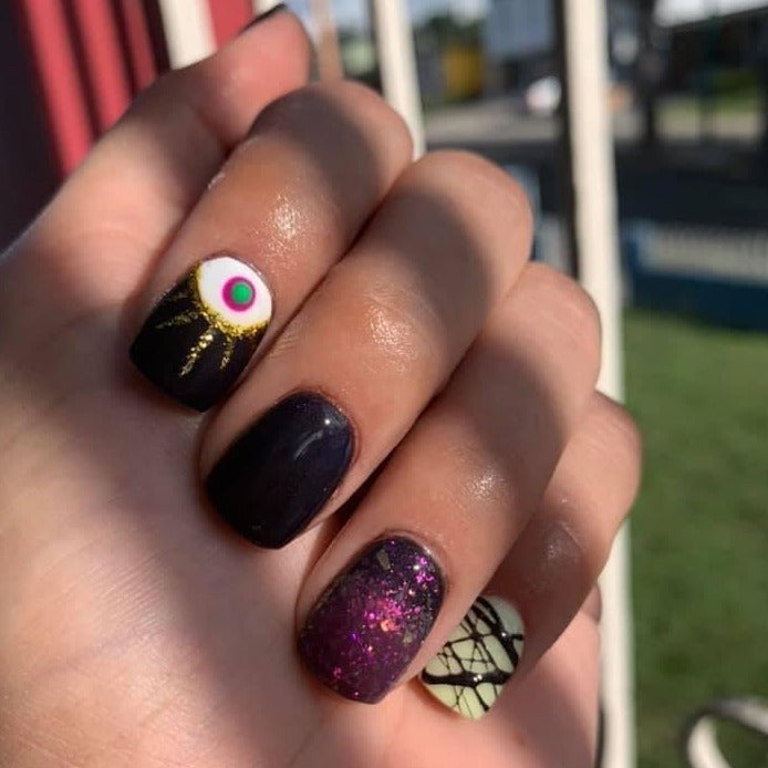 Hand with black glossy nails, one featuring a pink and white eye design, another with purple glitter, and one with black.