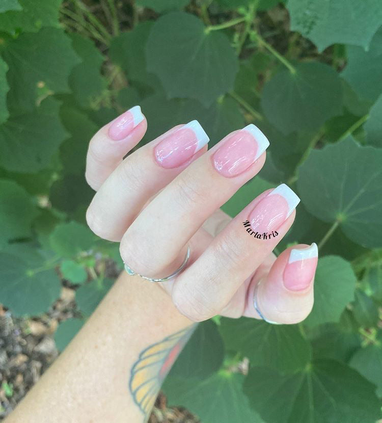 Hand with French tip pink dip powder nails and a small butterfly tattoo on the wrist against green leafy background.
