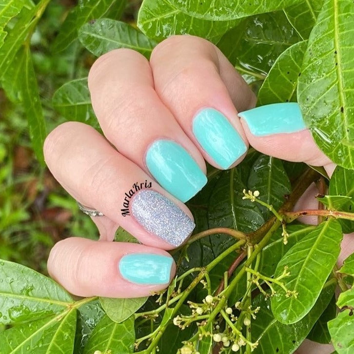 Hand with teal blue glossy nails and one glittery silver accent nail against green leaves background.