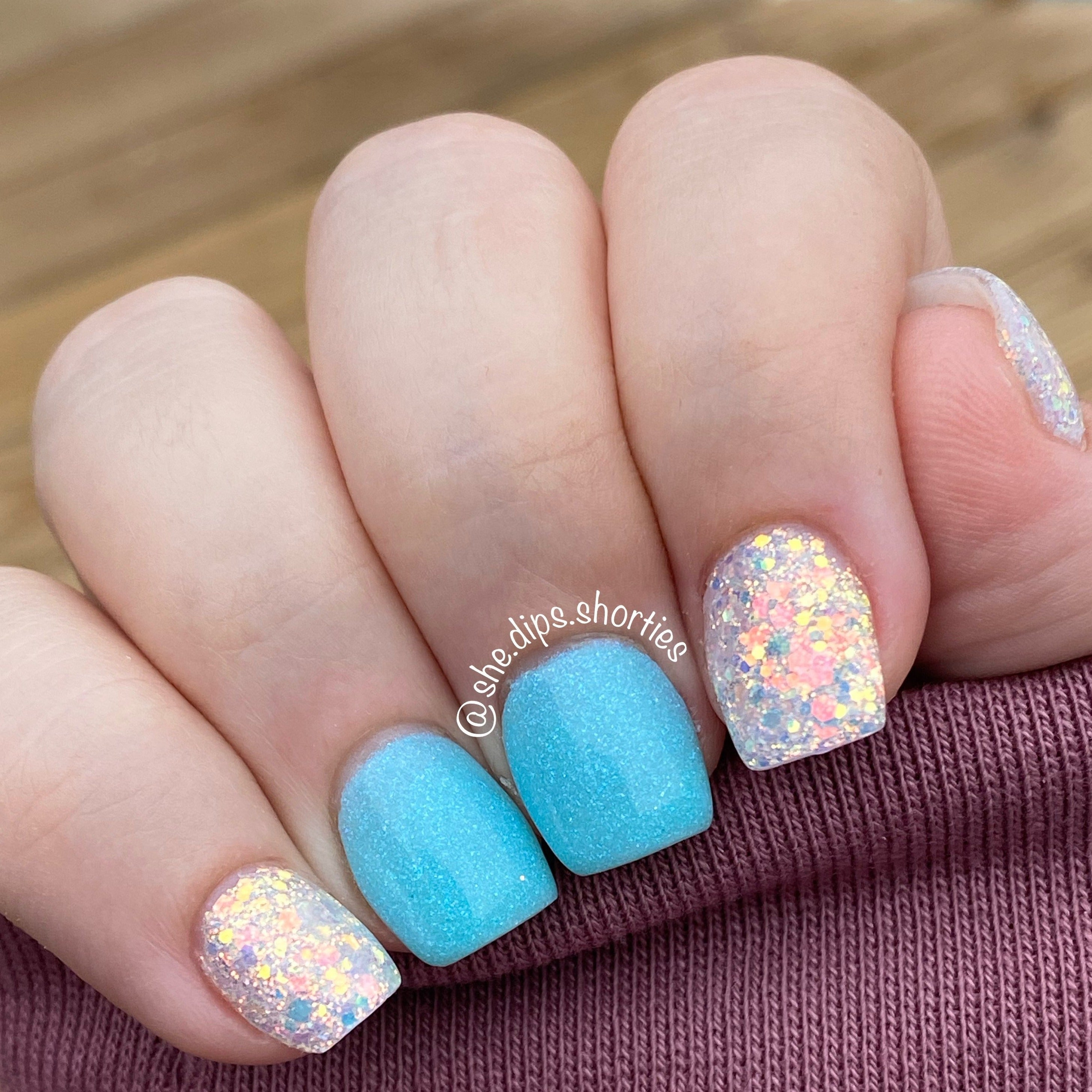 Short square nails with bright blue polish on two middle fingers and chunky iridescent white glitter on thumb, index, and.