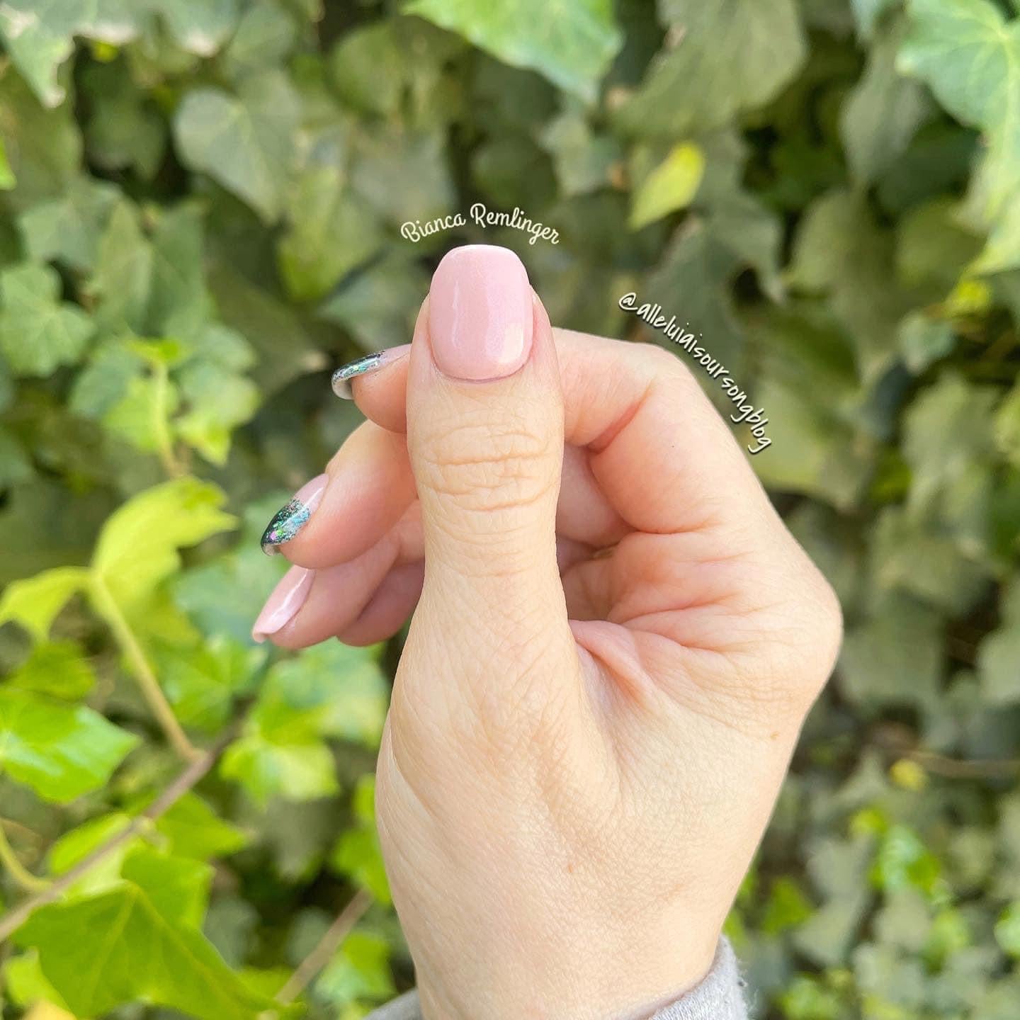 Hand with short, rounded nails painted in soft nude pink dip powder polish against green leafy background.