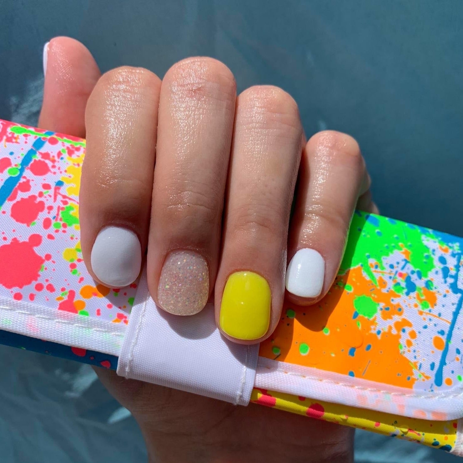 Hand with short nails featuring white, textured nude, and bright yellow polish holding colorful paint-splattered clutch.