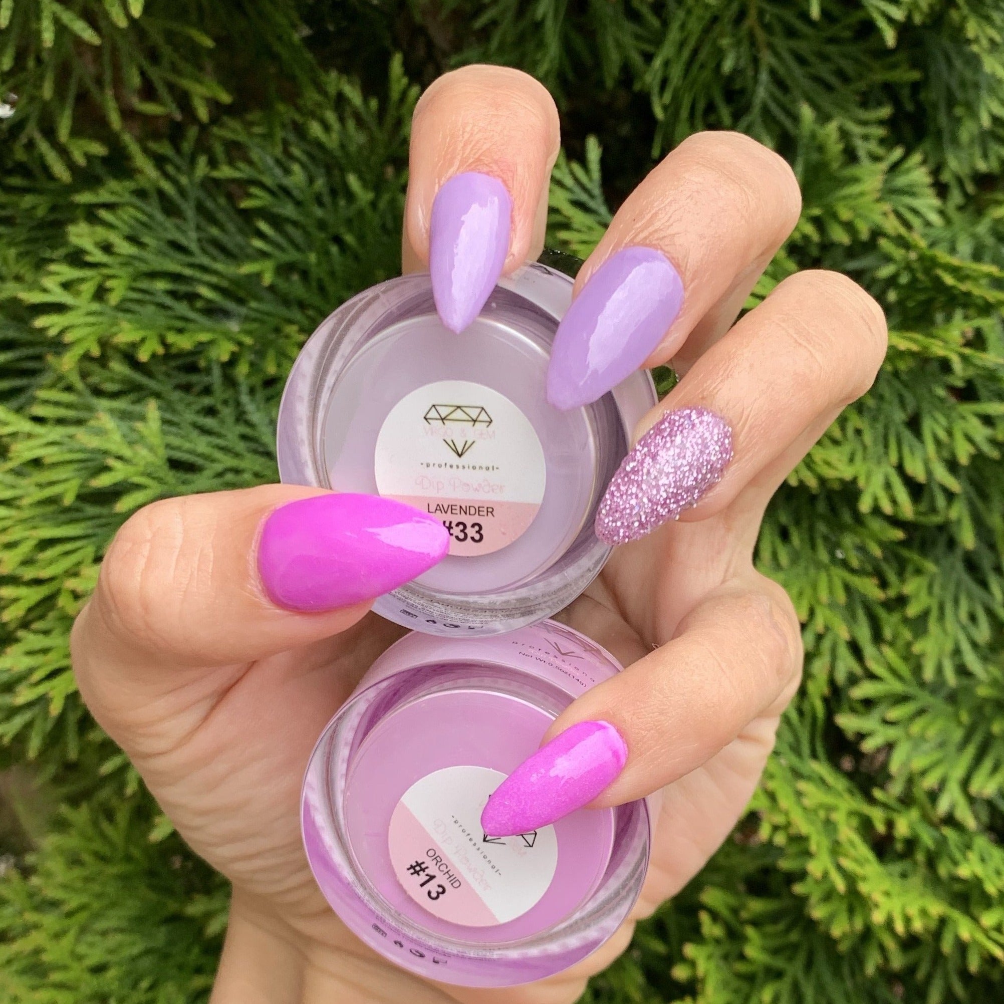 Hand with long almond nails in shades of purple and pink using solid dip powders, one nail with textured glitter finish,.