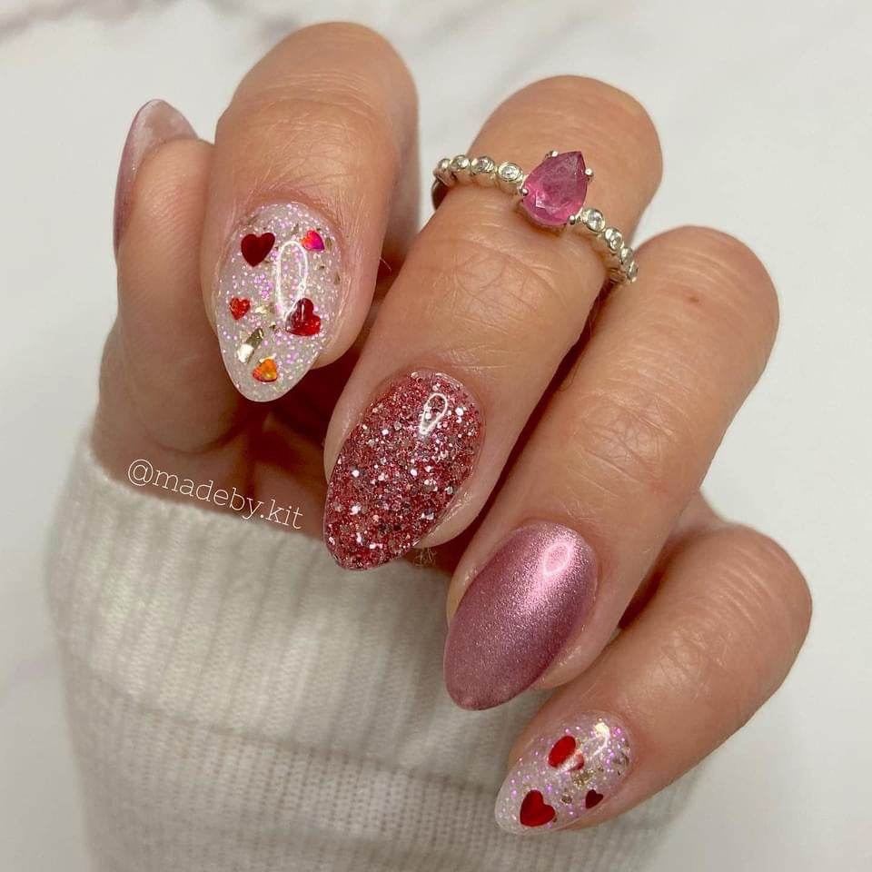 Hand with nails painted in glittery red, metallic pink, and clear polish with red heart accents.