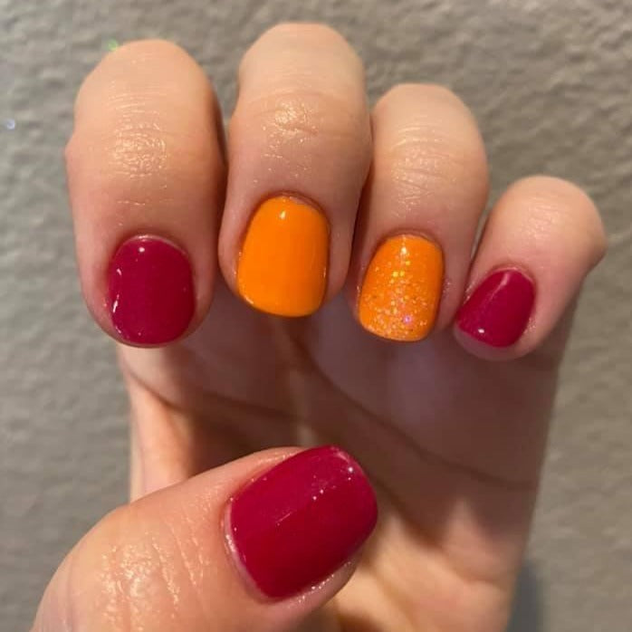 Hand with nails painted in deep red and bright orange, one orange nail features subtle glitter accents.
