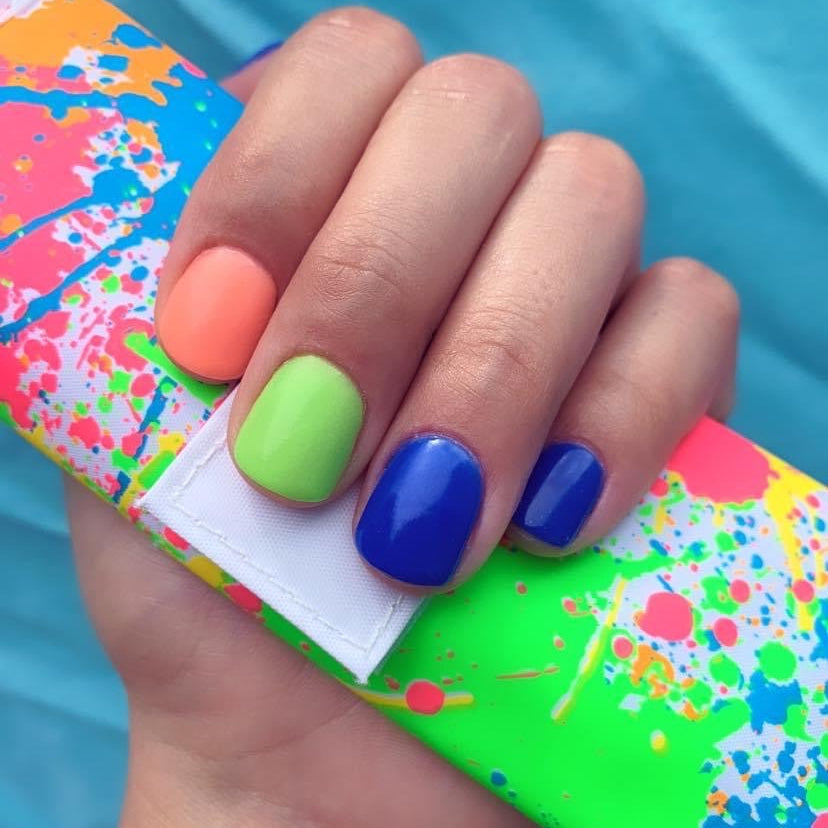 Fingernails painted with bright coral, lime green, and deep blue dip powder polish on colorful splattered clutch.