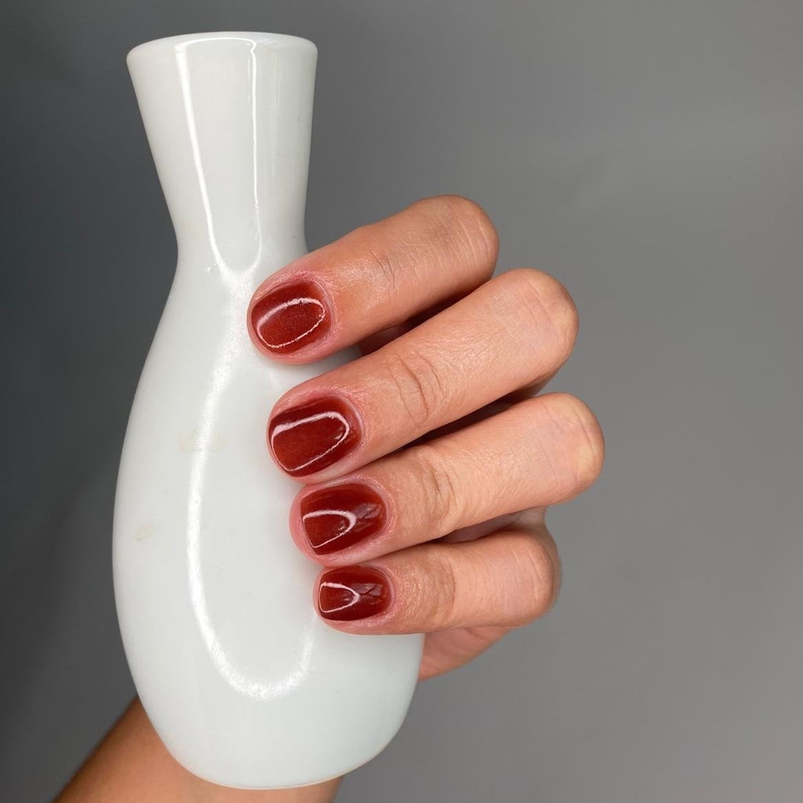 Hand with glossy deep brown-red gel nails holding a smooth white ceramic vase against gray background.