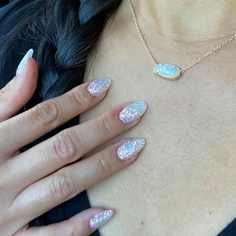 Fingernails with white iridescent fine glitter dip powder on almond-shaped nails against skin with gold necklace pendant.
