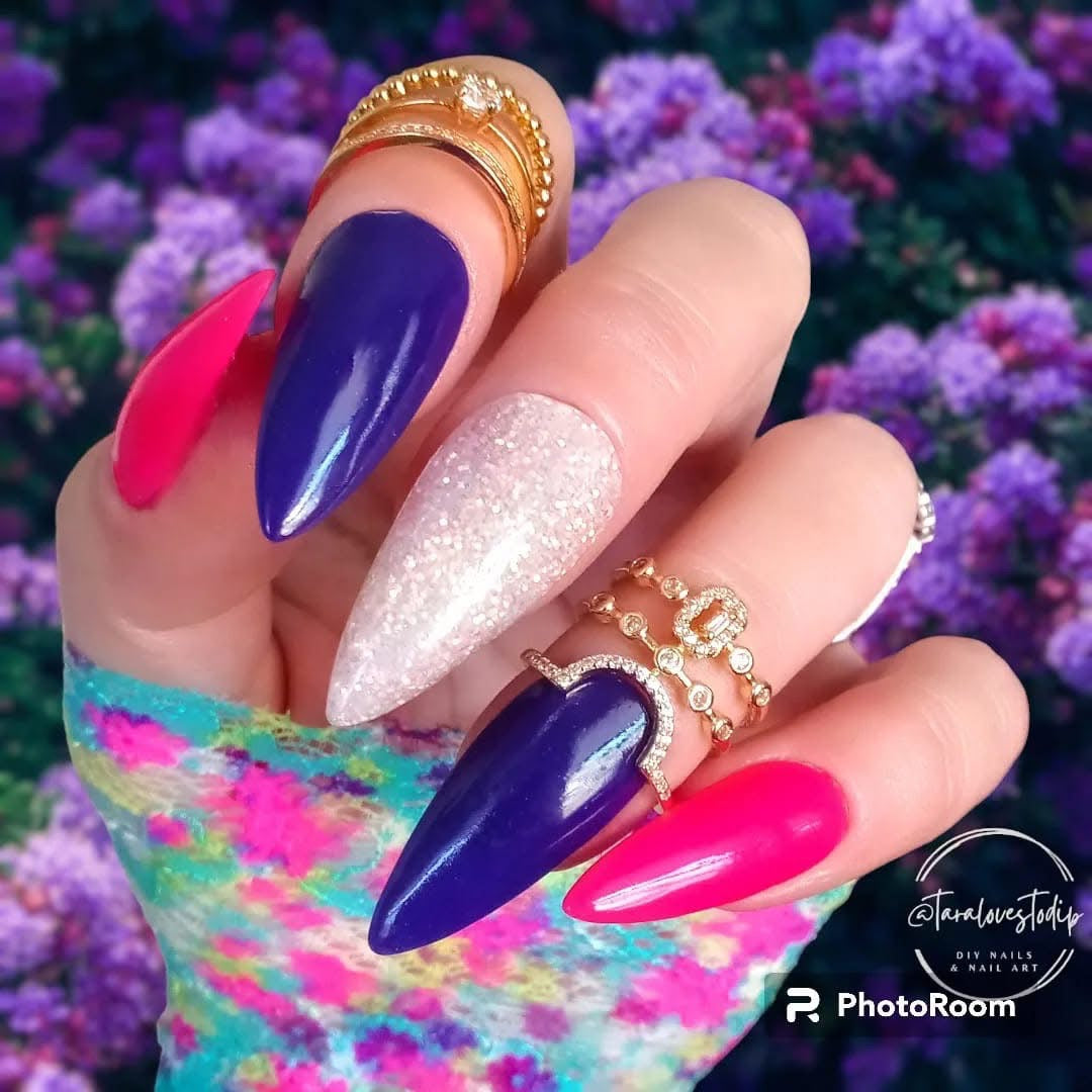 Long stiletto nails with dark purple, glittery white, and bright pink polish against floral background.
