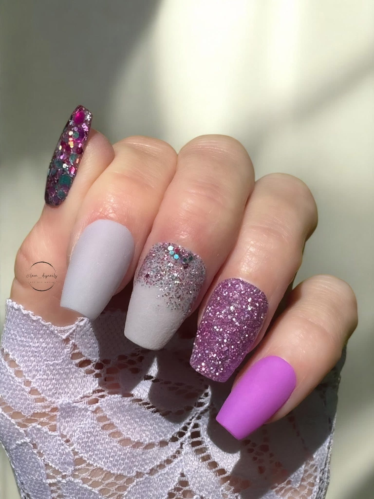 Hand with dip powder nails in shades of purple, featuring solid orchid, glitter, and rhinestone accents.