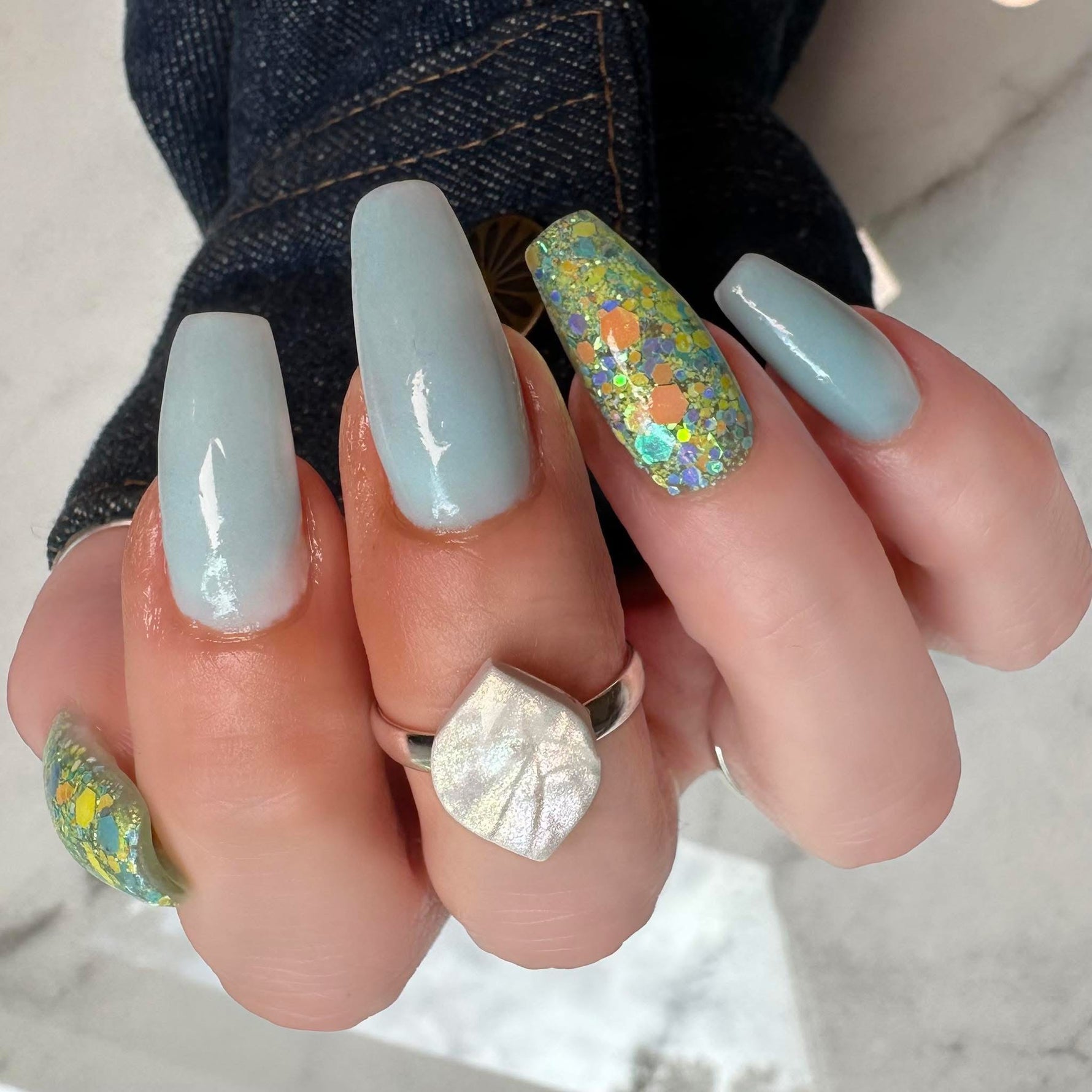 Long square nails with pastel blue polish and one accent nail featuring multicolor chunky glitter on a clear base.