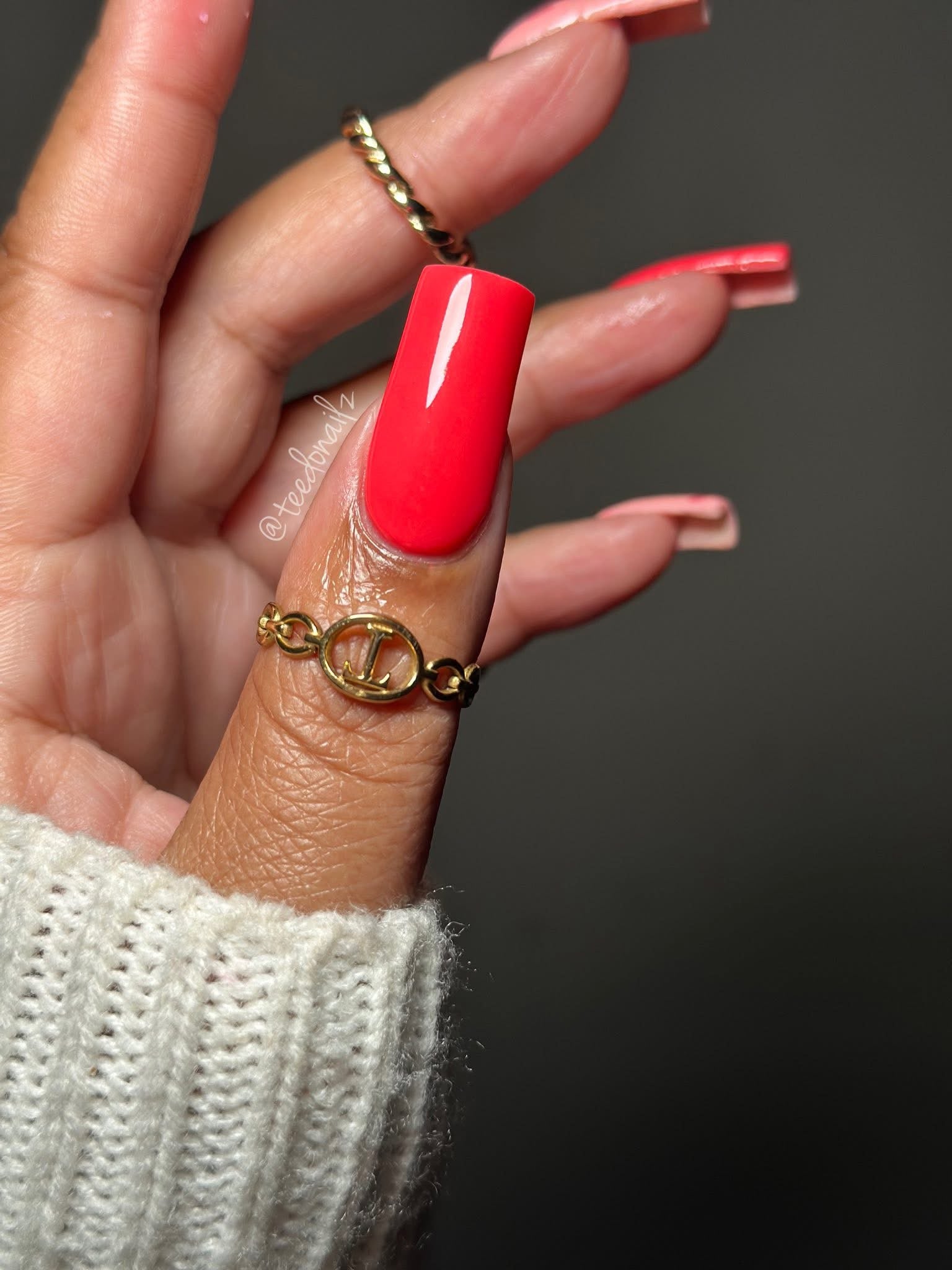Long square nails painted bright coral pink, glossy finish, gold rings on fingers, cream knit sleeve.