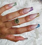 Hand with long coffin-shaped nails featuring fine glitter tips in green, purple, gold, and blue over nude base polish,.