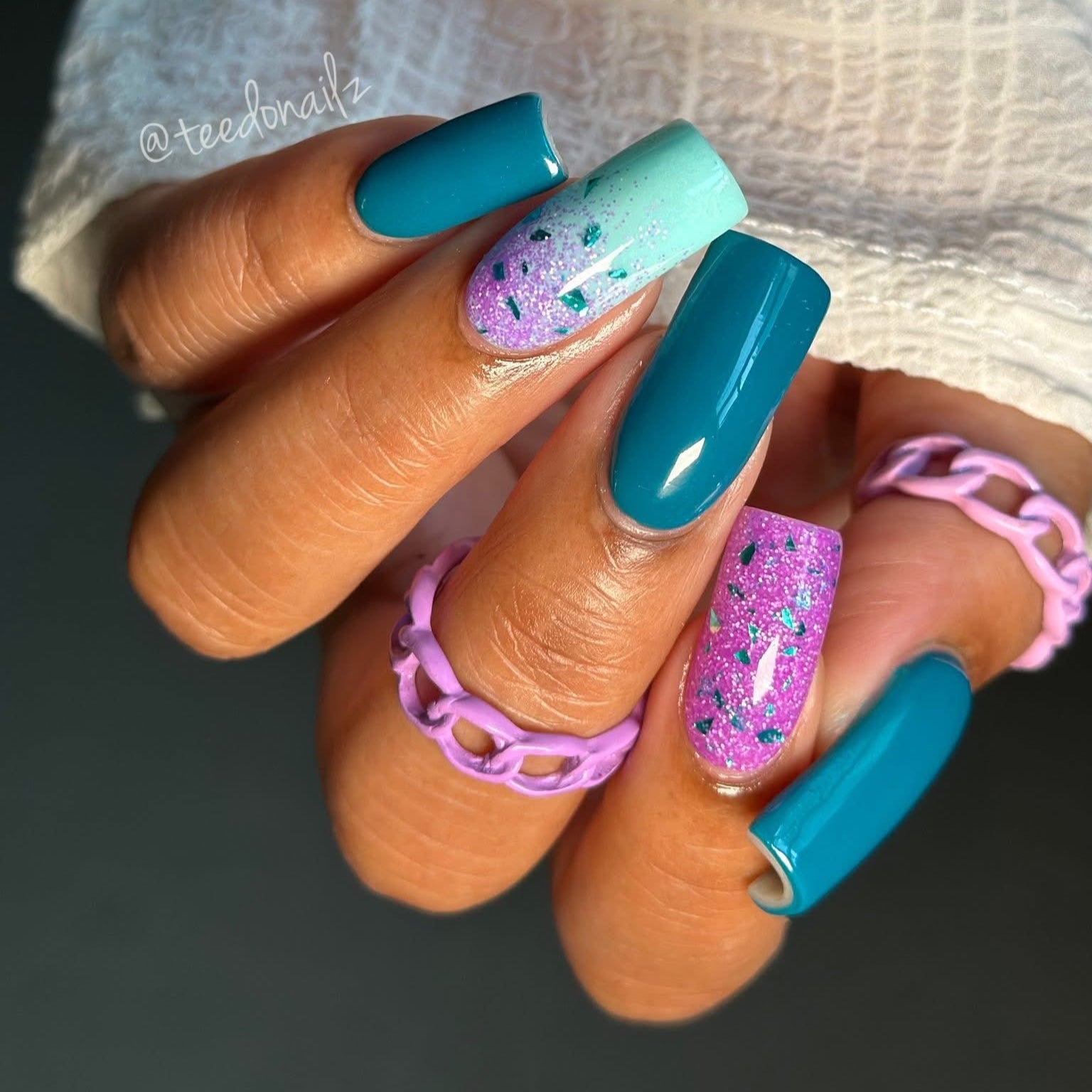 Hand with teal and multicolored glittery nails wearing a white textured sleeve.