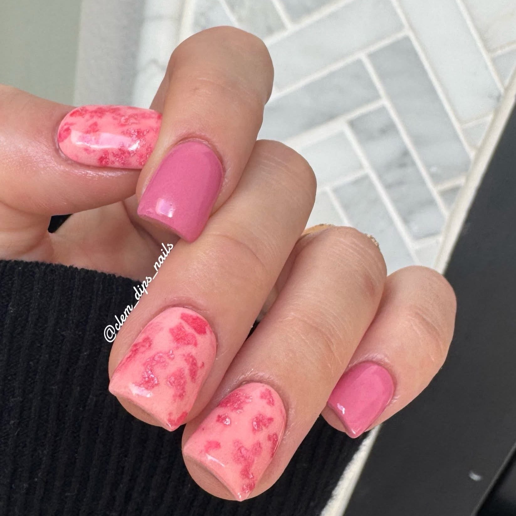 Close-up of a hand with peach dip powder and pink foil on a tiled floor background.