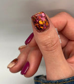 Chunky glitter dip powder on thumbnail with orange, pink, and cranberry hues and glossy finish.