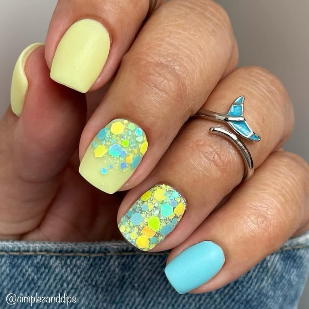 Matte pistachio green nails with yellow and blue hex glitter accent nails and silver butterfly ring on finger.