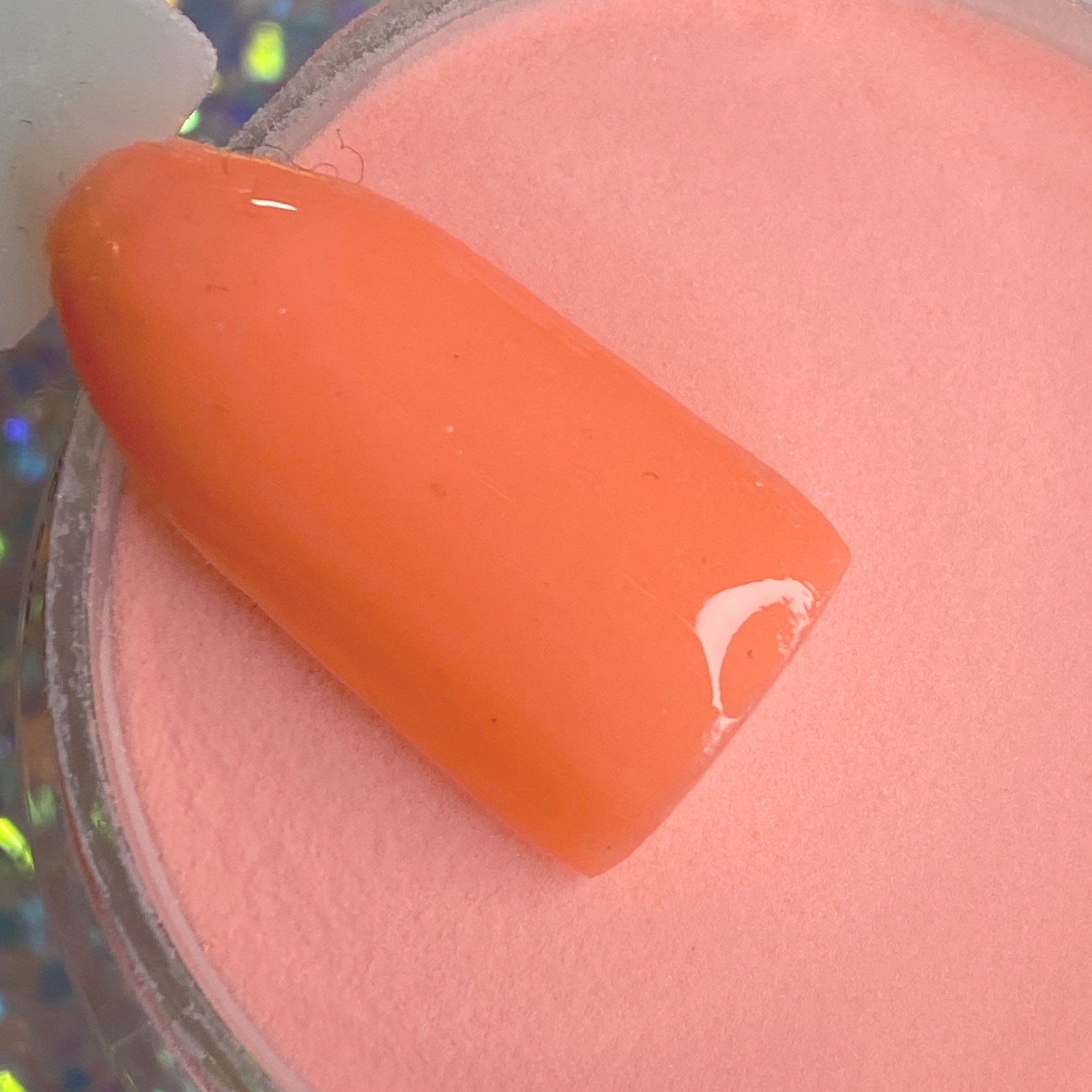 Glossy pastel coral-orange dip powder on a nail tip over matching powder jar.