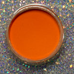 Bright solid orange dip powder in clear round jar on sparkling holographic surface.