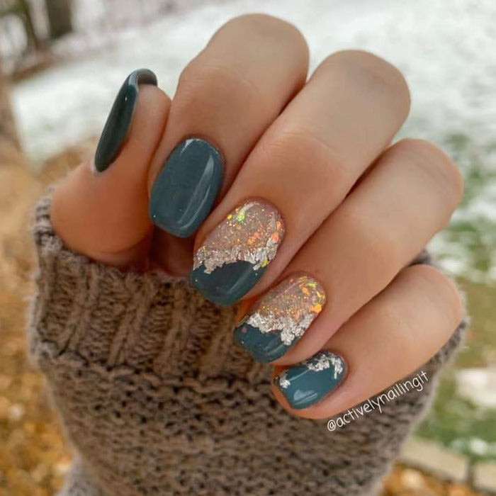 Hand with blue-gray matte nails and accent nails featuring gold foil and white floral designs.