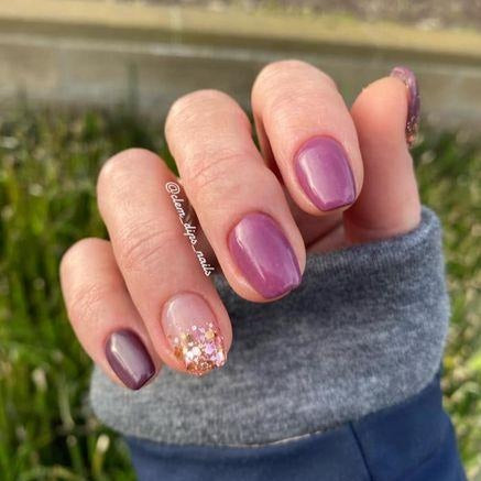 Hand with purple dip powder nails, one nail with gold and pink glitter accents, wearing gray and navy sleeve.