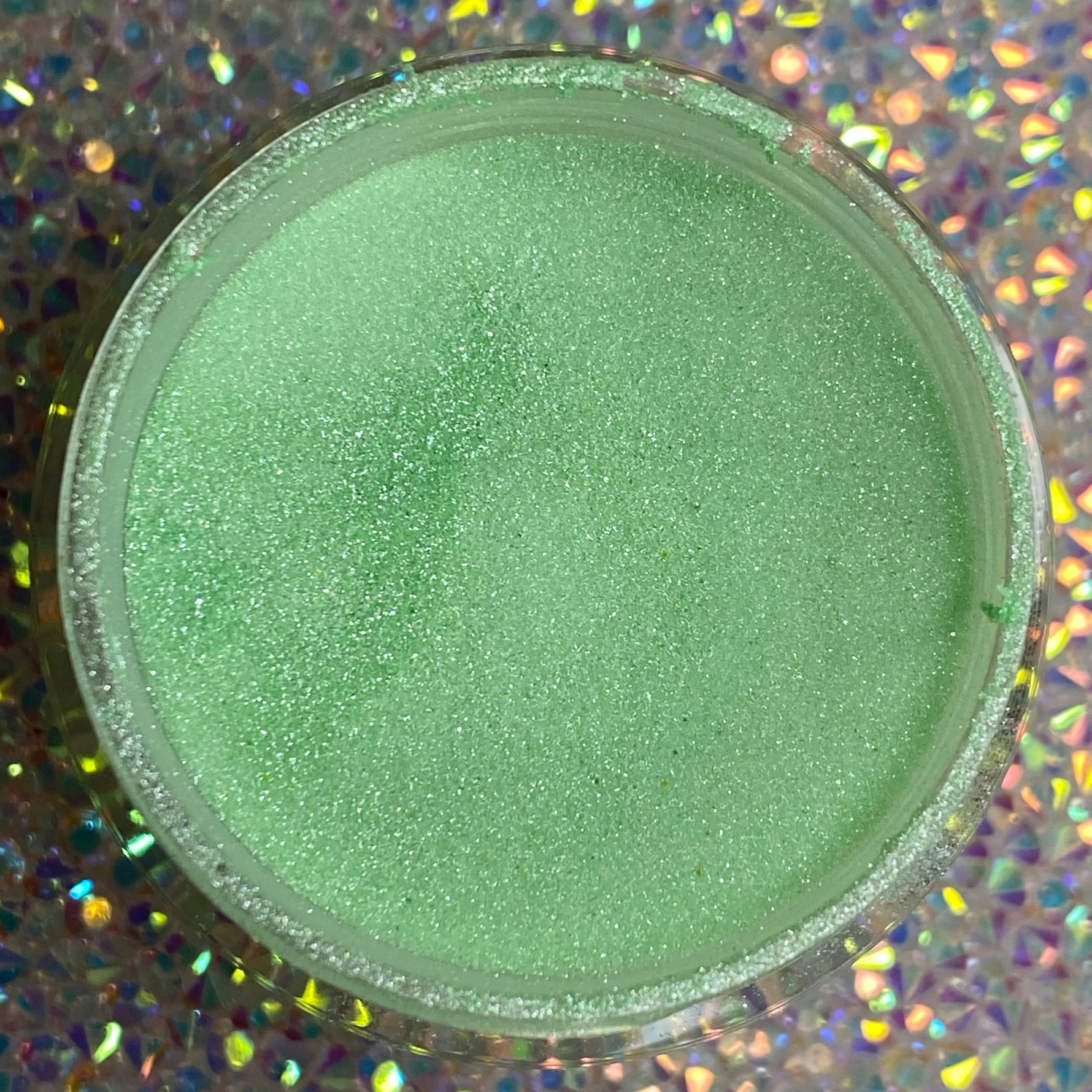 Open jar of fine mint green glitter dip powder with subtle shimmer on holographic background.