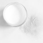Clear fine dip powder in a small round jar with some powder spilled next to it on a white surface.