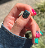 Short nails with chunky green and multicolor glitter on the thumb, bright pink on the index and middle fingers, and teal on.