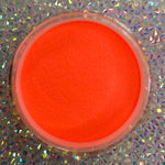Bright neon orange glow-in-the-dark dip powder in a round clear container on a holographic surface.