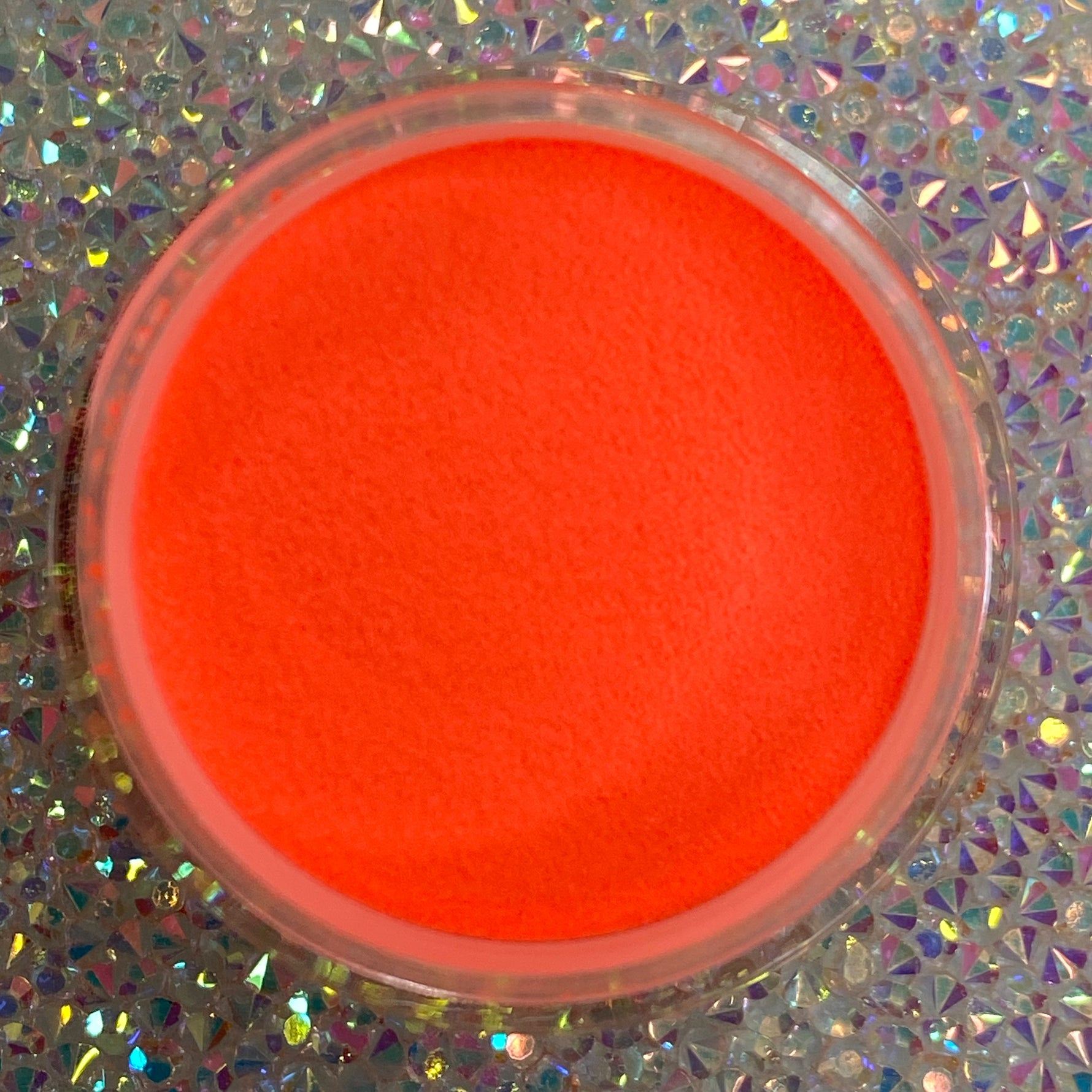 Bright neon orange glow-in-the-dark dip powder in a round clear container on a holographic surface.