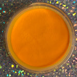 Bright neon orange glow-in-the-dark dip powder in a round container against a holographic background.