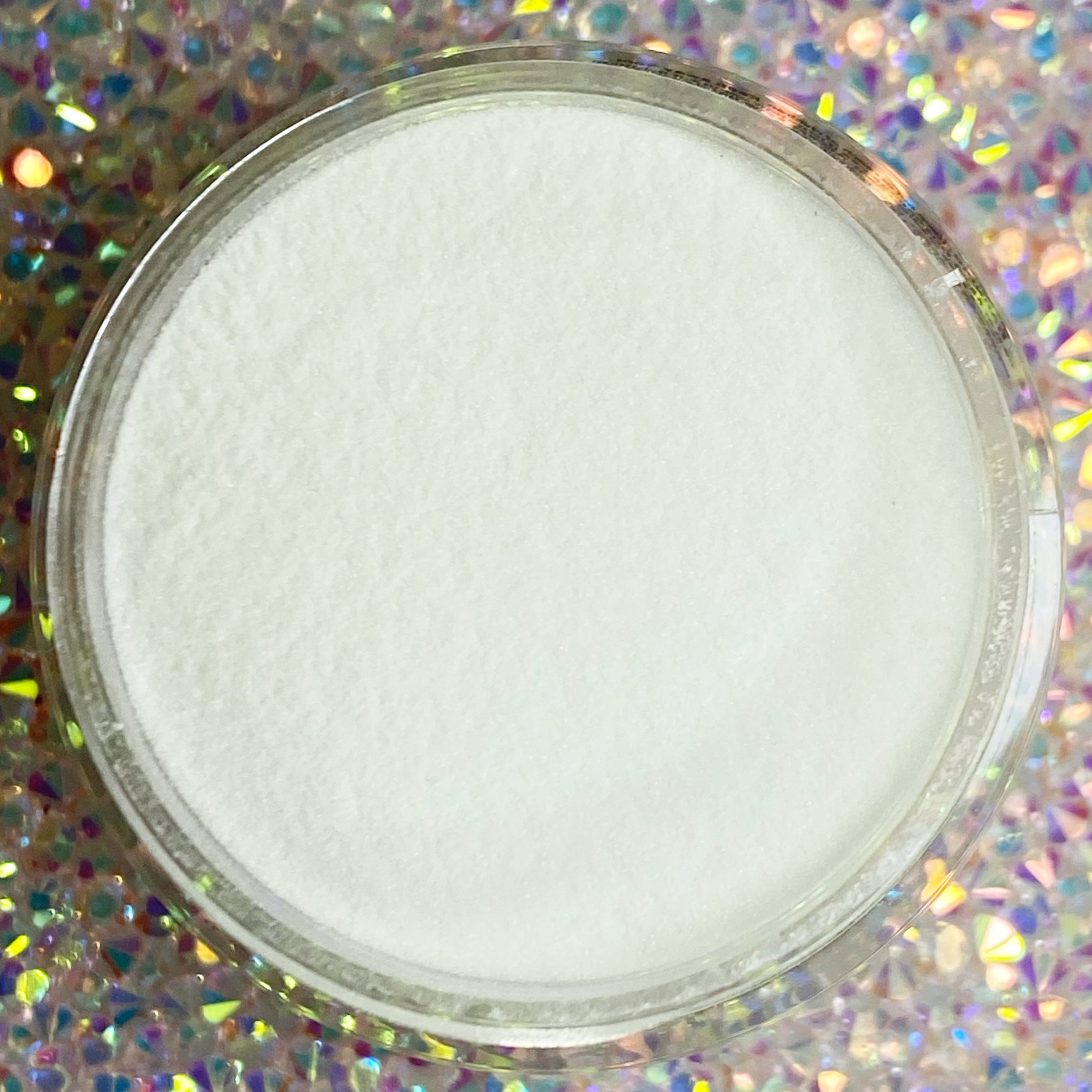 Milky white dip powder in clear round container on sparkly holographic background.