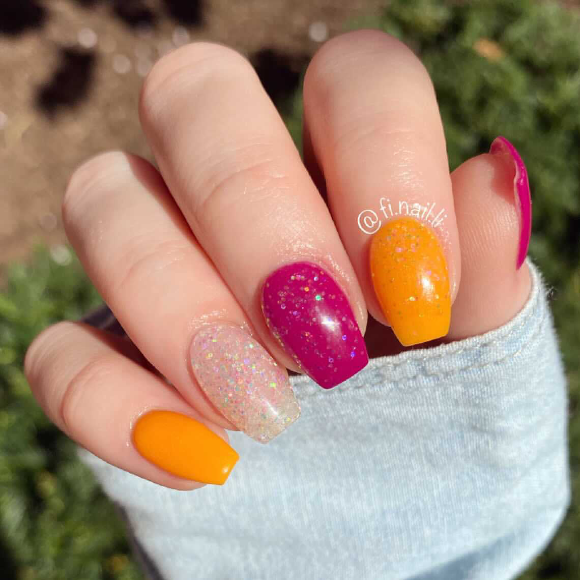 Hand with orange, pink glitter, and clear nails with pink flakes, outdoors in sunlight.