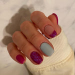 Hand with nails painted in matte dusty blue, glossy magenta, and glittery purple accents on a marble background.